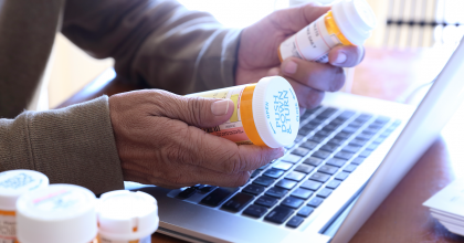 Photo of senior citizen reading labels on prescription drug bottles while at his laptop computer.